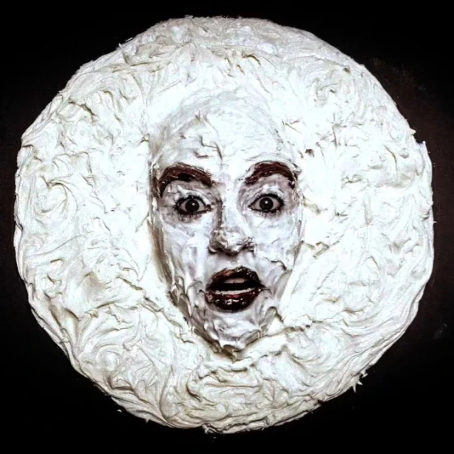 A dramatic portrait features an adult female with white cream covering her face and hair, set against a stark black background.