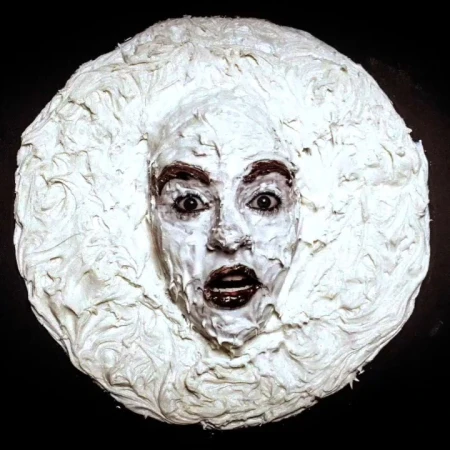 A dramatic portrait features an adult female with white cream covering her face and hair, set against a stark black background.
