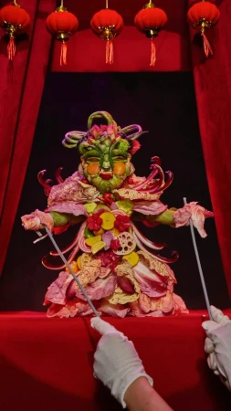 Vegetable puppet on stage with red curtains and hanging lanterns, controlled by hands in white gloves holding rods.