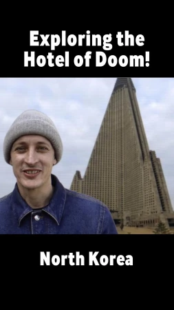 Man in beanie and denim jacket stands in front of Ryugyong Hotel, North Korea. Tall, pyramid-shaped building. Cloudy sky backdrop.
