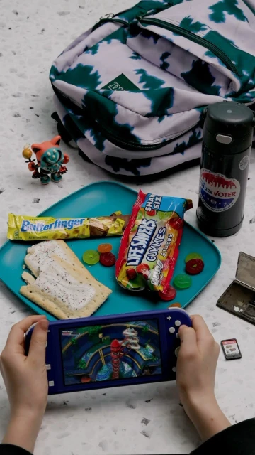 Child plays a video game on a handheld console. A backpack, snacks, and a water bottle are set on a speckled table surface.