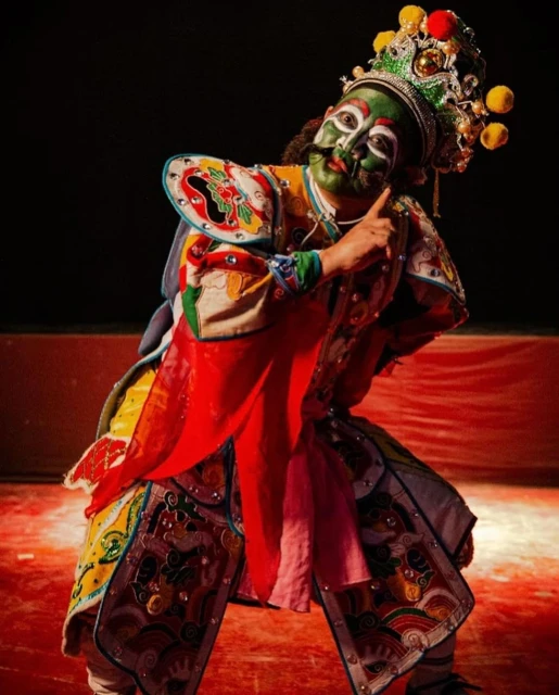 Chinese opera performer in costume and face paint on a red stage. He is making a hand gesture and wearing a decorative headdress.