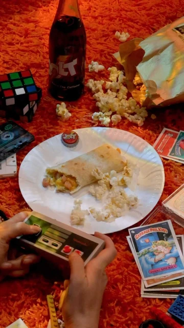 Overhead shot of hands playing a retro game, with a burrito, drink, and snack on orange shag rug.