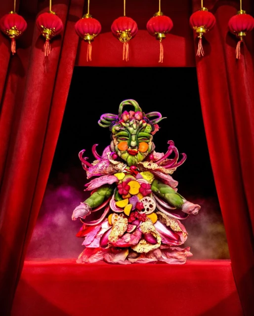 Festive food art creation with vegetables and fruits in a studio setting. Red curtains and decorative Chinese lanterns provide a colorful backdrop.