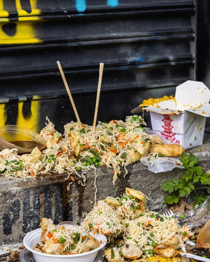 Close-up of discarded Chinese food, including noodles and takeout containers, spills onto a concrete city sidewalk with spray paint.