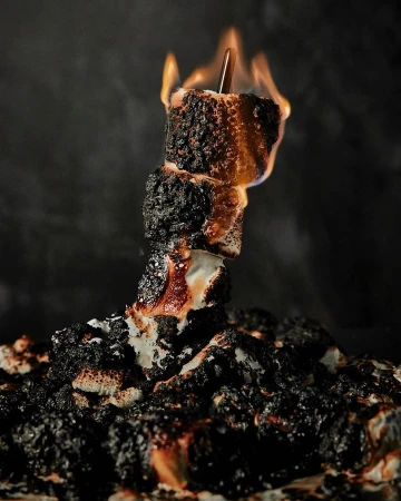 Stacked marshmallows on a skewer, intentionally charred and engulfed in small flames against a dark background, creating a dramatic food shot.