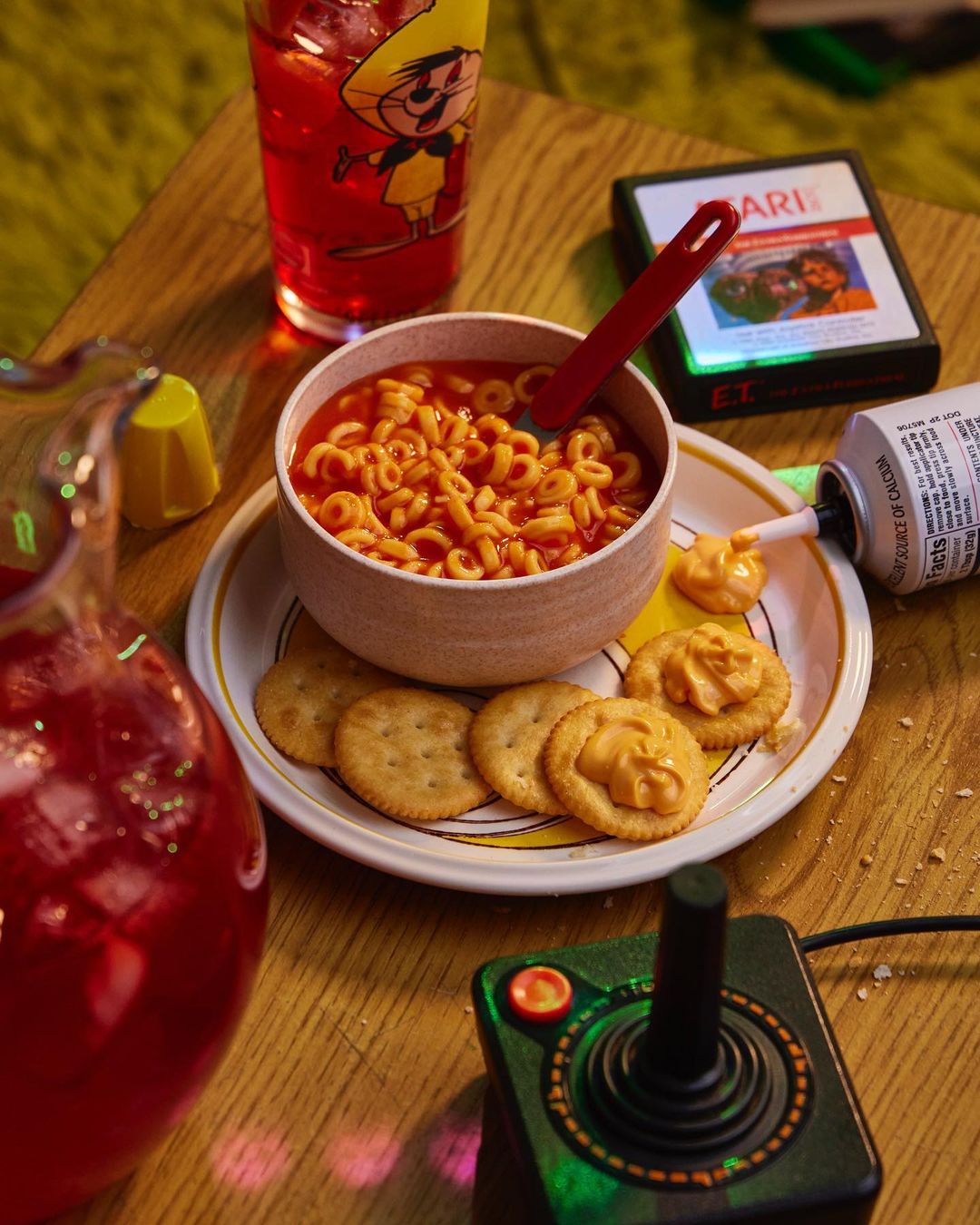 Retro food and gaming scene on a wood table featuring alphabet soup, crackers with cheese spread, a joystick, and a juice pitcher.