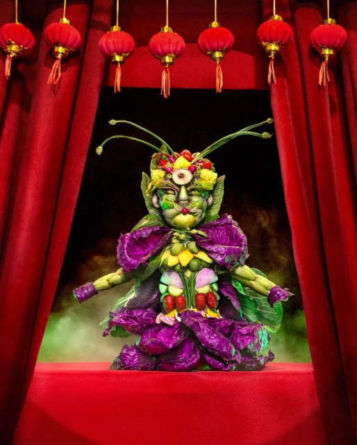 Costumed performer on a red stage in a full, centered shot. The costume is constructed of colorful vegetables and fruits. Lanterns above.