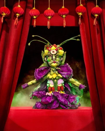 Costumed performer on a red stage in a full, centered shot. The costume is constructed of colorful vegetables and fruits. Lanterns above.