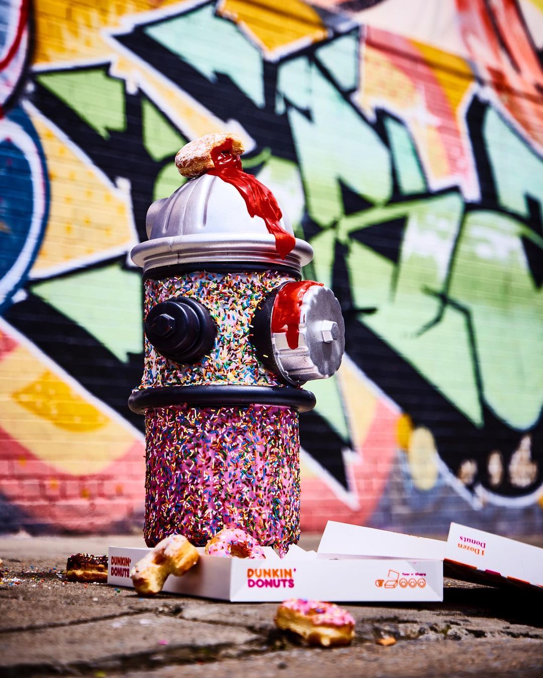 A fire hydrant art installation covered in sprinkles and donuts, staged on a city street with graffiti, and Dunkin' Donuts packaging.