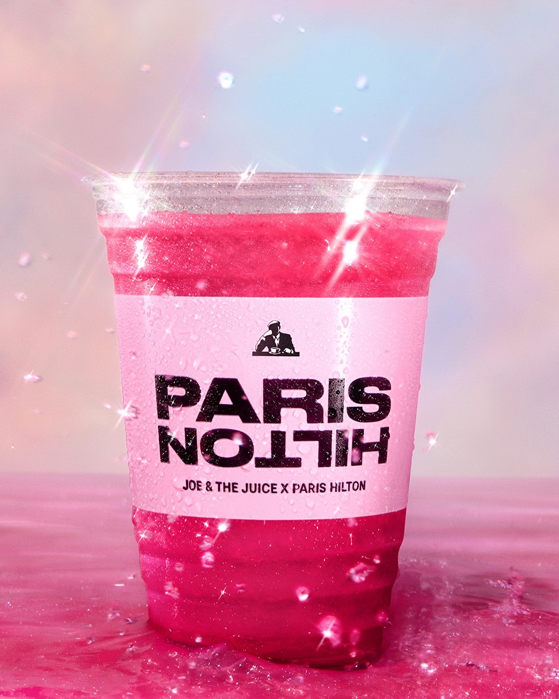 Close-up of a bright pink Paris Hilton Joe & The Juice drink in a clear plastic cup, pink surface, and a dreamy sky background.