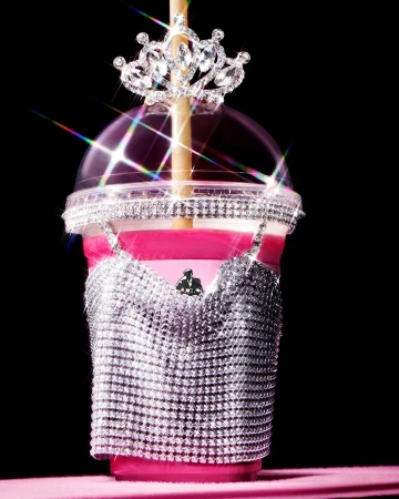 Novelty drink in a clear plastic cup with a straw, sparkly bra-shaped decoration, and a tiara against a dark background.