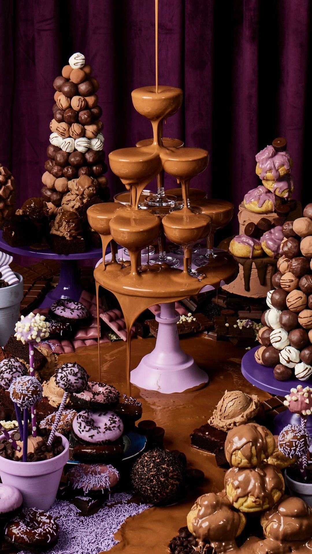 Extravagant chocolate dessert table featuring towers of chocolate truffles, glazed donuts, and champagne glasses filled with chocolate sauce.