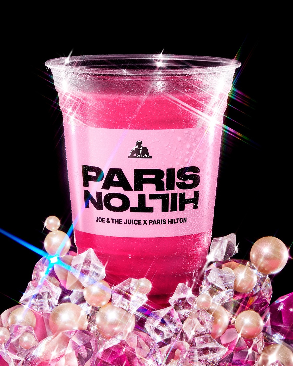 Studio shot of a pink-colored beverage in a plastic cup with ice and pearls, prominently featuring the logo of Paris Hilton.