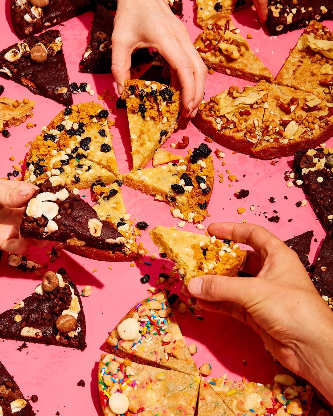 Overhead shot features hands reaching for several slices of dessert pizzas with various toppings on a vivid pink surface.
