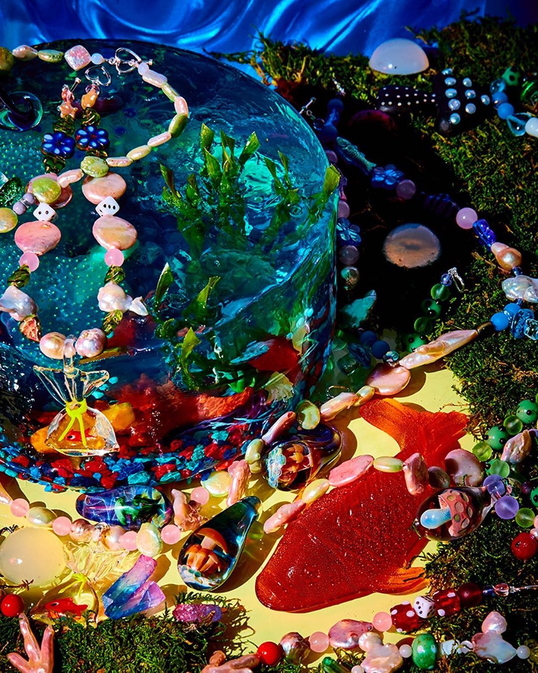 Whimsical still life featuring a clear, spherical container holding abstract blue and green shapes, surrounded by colorful beads and mossy texture.