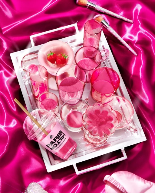 Still life arrangement of pink-themed drink, desserts, makeup brushes, and accessories on a silver tray against a pink satin background.