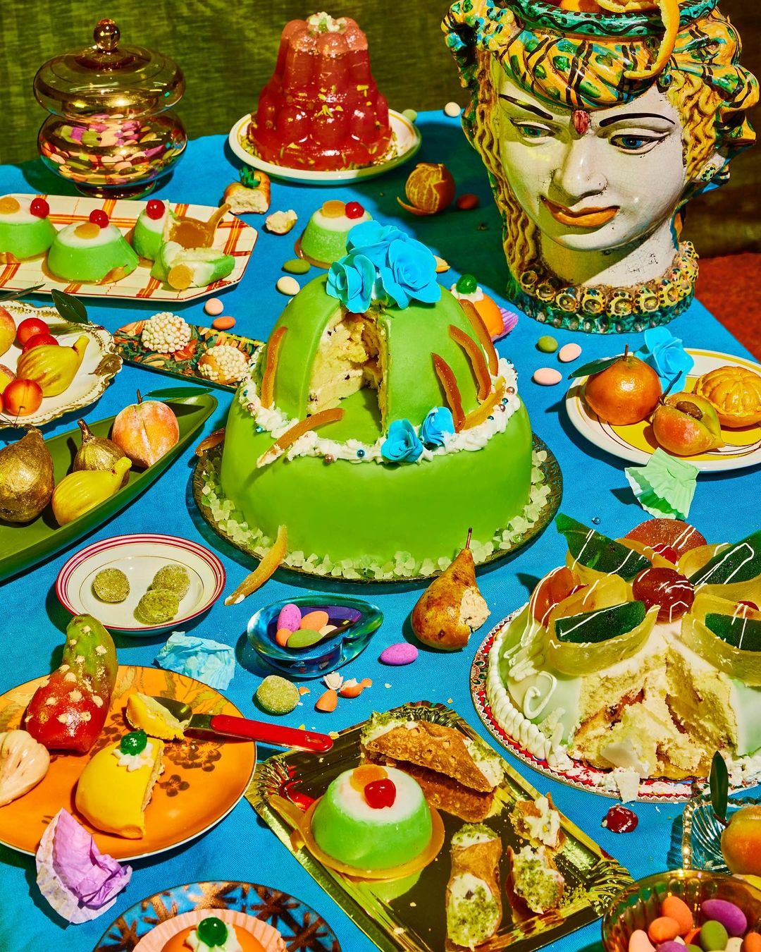 A vibrant still life showcases a variety of cakes, candied fruits, and desserts arranged on a turquoise tablecloth, evoking a sense of abundance.
