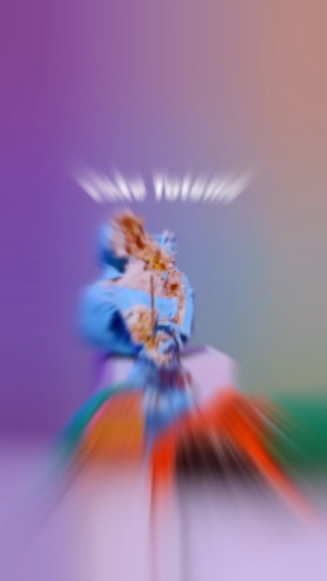 Blurred image of a person with blue and brown clothing sitting on a pink and white surface with a purple and orange background.