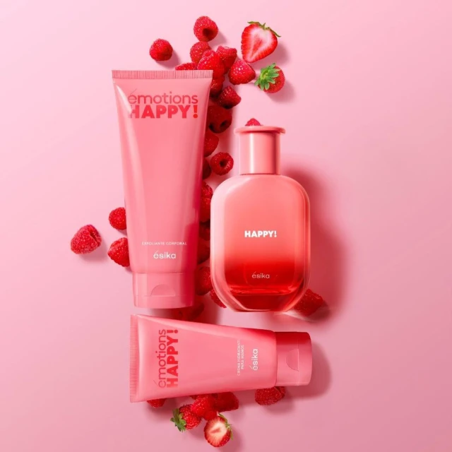 Still life of pink 'Esika Emotions Happy!' fragrance bottle and body lotion tubes surrounded by raspberries and cut strawberry on pink.