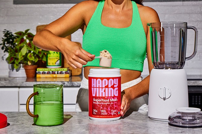 Adult female adding protein powder to a blender, wearing a green sports bra in a kitchen setting with countertop containers.