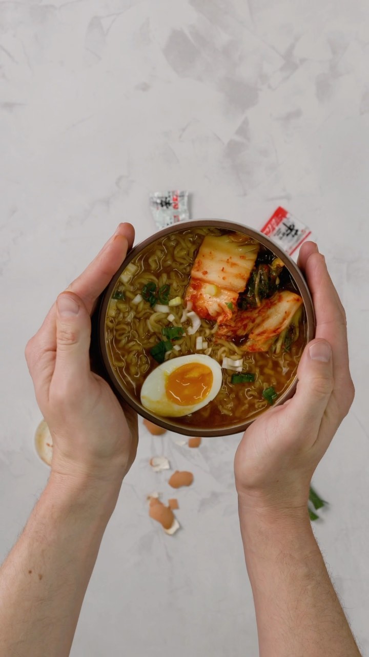 Overhead shot of a person holding a bowl of ramen with kimchi, soft boiled egg, and scallions, with spilled eggshells on a white surface.