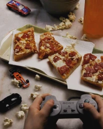 Person holding a video game controller next to a tray of pizza slices, scattered popcorn, and toy cars.
