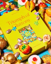 A yellow puzzle box titled Triumph of Gluttony surrounded by colorful puzzle pieces, dried figs, pastries, and metallic candies.