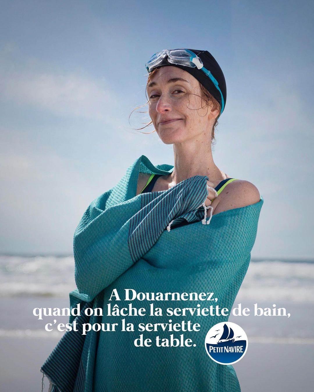 Woman in swim cap and goggles wrapped in a blue towel on a sunny beach, smiling slightly. Clear blue sky and sea in background.