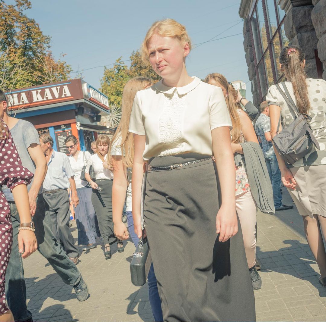 Woman in white top and black skirt walks in a crowd on a city street next to a small vendor kiosk on a sunny day.