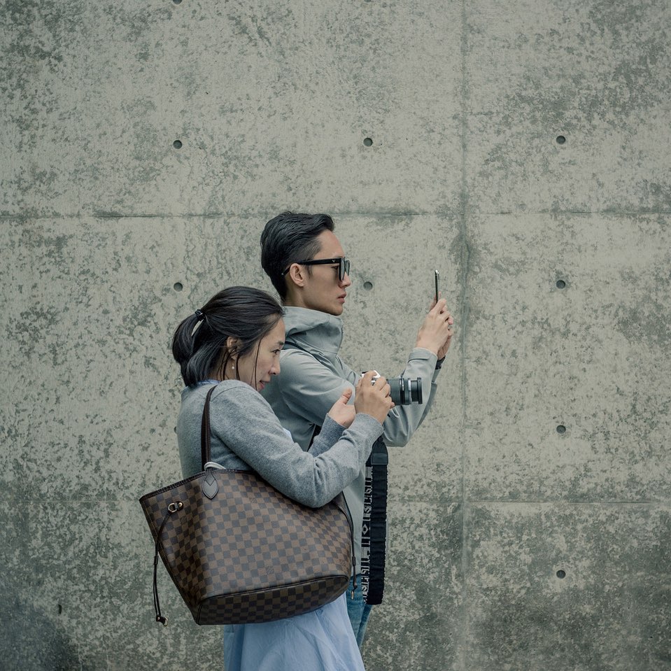 A couple stands against a concrete wall with the man holding a smartphone and the woman holding a digital camera; both are taking pictures.