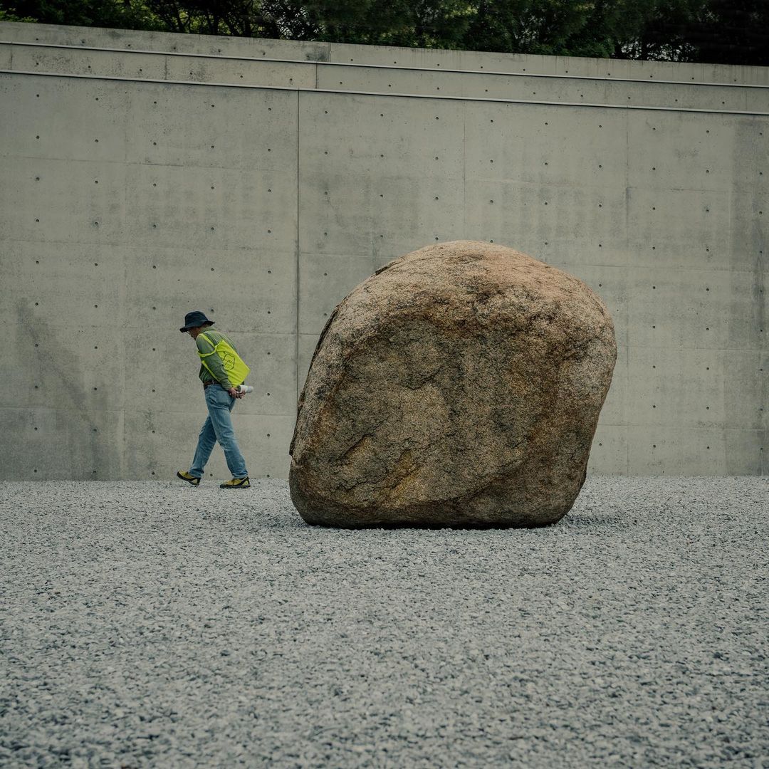 Person walking past a large boulder on a gravel surface against a concrete wall backdrop. Natural light and simple composition.