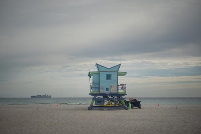 Miami Beach lifeguard station under cloudy sky with cargo ship. Aqua shelter, green accents, beach sand, distant ship, and cloudy sky.