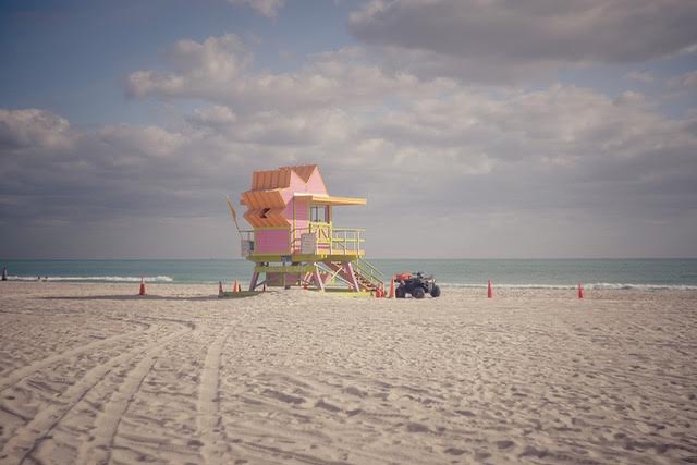 Miami Beach lifeguard stand in pink and yellow, on a sunny day with an ATV nearby, set against a backdrop of the ocean and cloudy sky.
