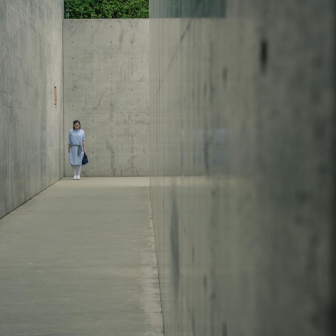 A young woman in a light-blue dress stands in a concrete hallway with an ambiguous gaze, set against a muted, minimalist backdrop.