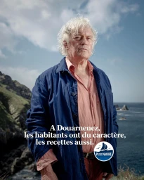 Elderly man with white hair wearing a blue jacket and pink shirt standing outdoors near a rocky coast.