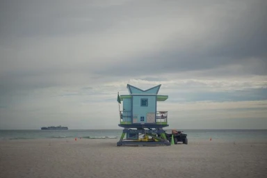 Blue and green lifeguard tower on a wide sandy beach under an overcast sky with a cargo ship on the horizon.