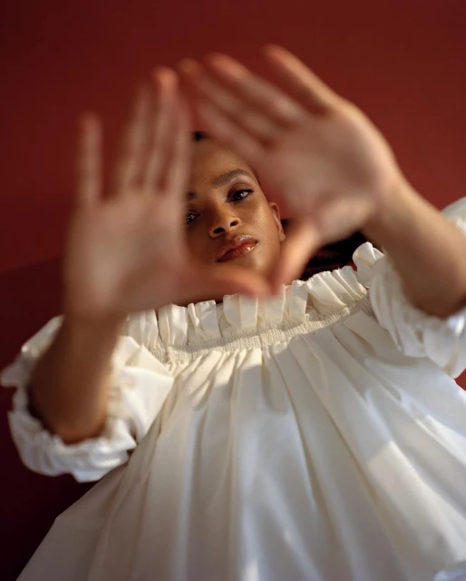 Portrait of a young adult woman in a white dress making a triangle with her hands indoors against a red background, from a low angle.