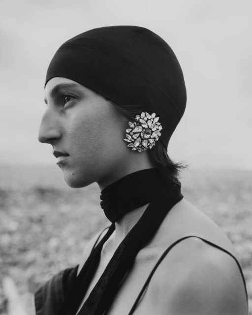 Monochromatic portrait of a woman with a black head covering and large floral earring, gazing left against a blurred neutral background.