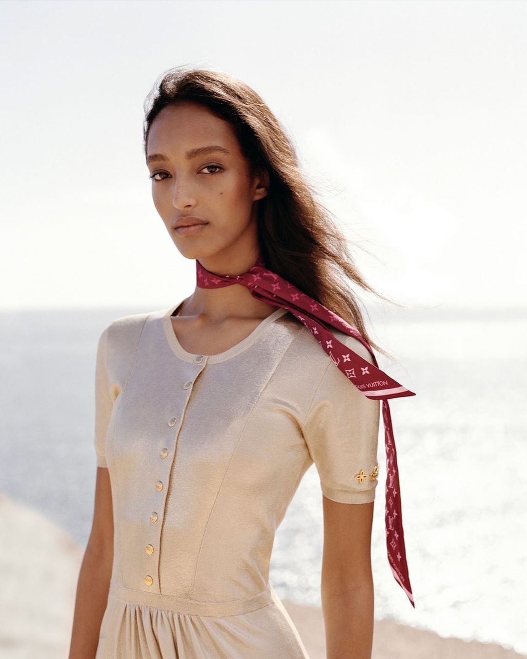 An adult female model poses outdoors wearing a light-beige dress and a burgundy silk scarf with a Louis Vuitton logo pattern.