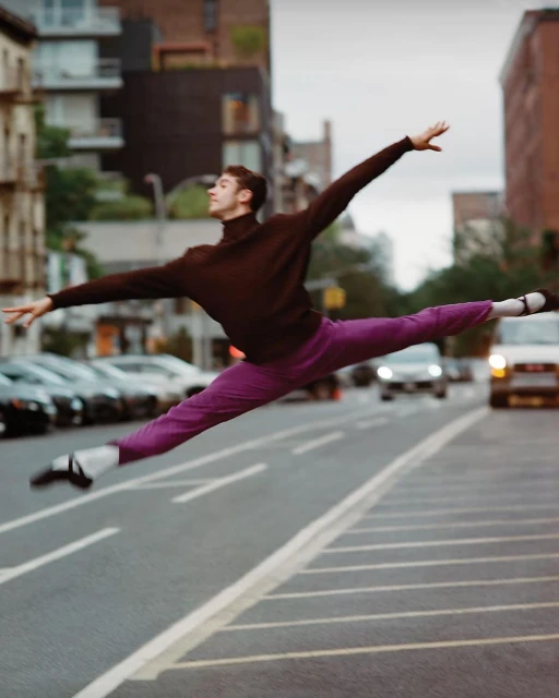 Male ballet dancer leaps mid-air on city street in purple pants, brown shirt, and ballet shoes, with urban buildings and cars in background.