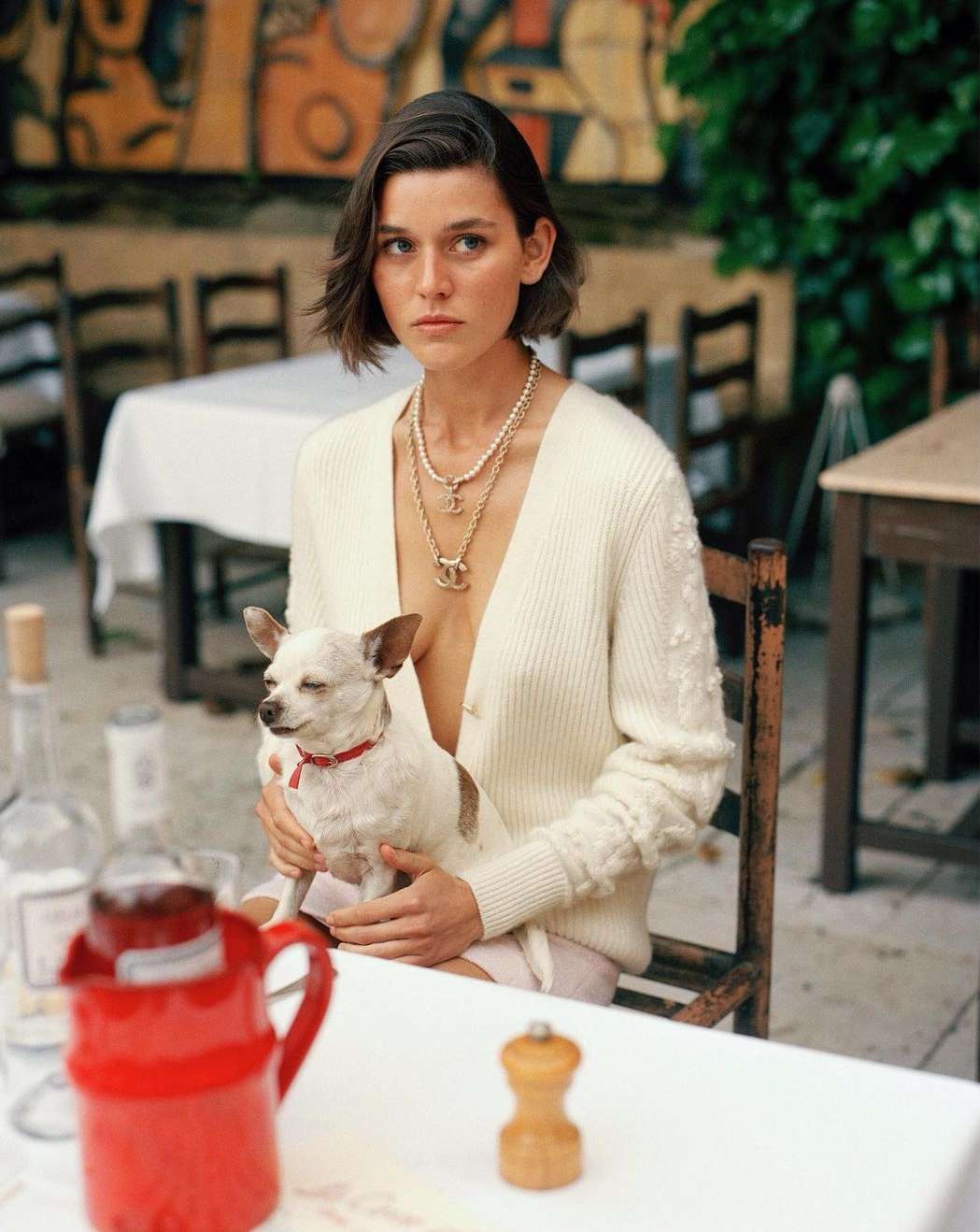 Elegant woman in cardigan seated with chihuahua in outdoor cafe, adorned with layered necklaces. Neutral palette, natural light.