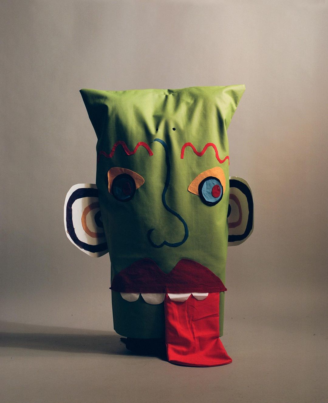 Playful green fabric sculpture with abstract facial features, including colorful stitched details and a protruding red tongue, set against a grey backdrop.