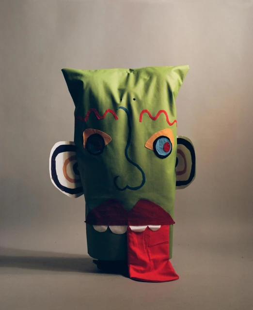 Playful green fabric sculpture with abstract facial features, including colorful stitched details and a protruding red tongue, set against a grey backdrop.