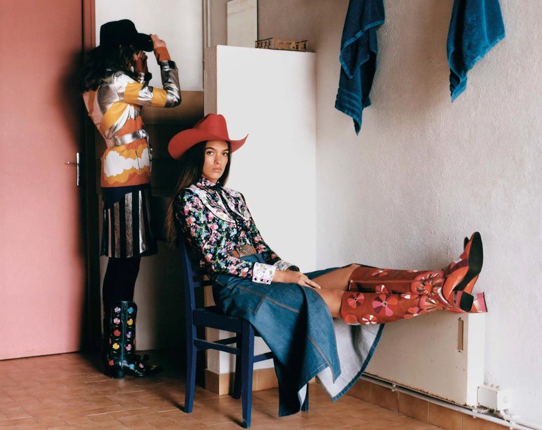 Two women pose in a room with pink door and vintage fashion, one seated on a blue chair, the other with binoculars.