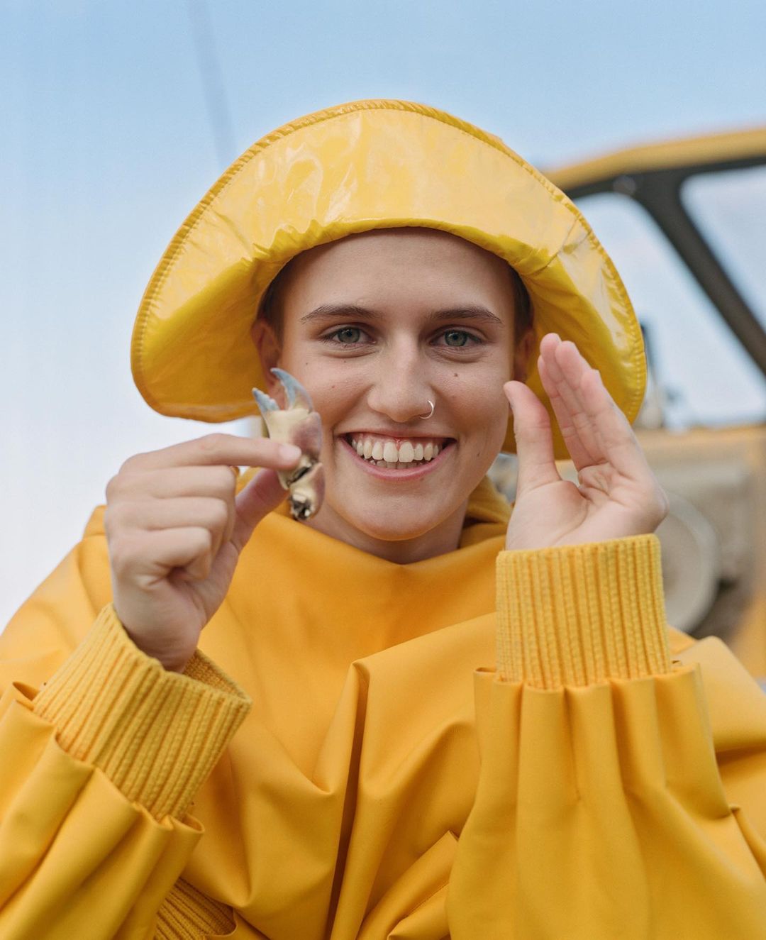 Smiling woman in yellow fishermans raincoat and hat holds crab claw outdoors by the water; blue sky visible above. 