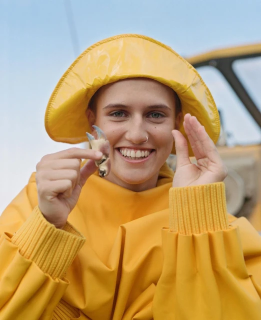 Smiling woman in yellow fishermans raincoat and hat holds crab claw outdoors by the water; blue sky visible above. 