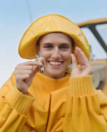 Smiling woman in yellow fishermans raincoat and hat holds crab claw outdoors by the water; blue sky visible above. 