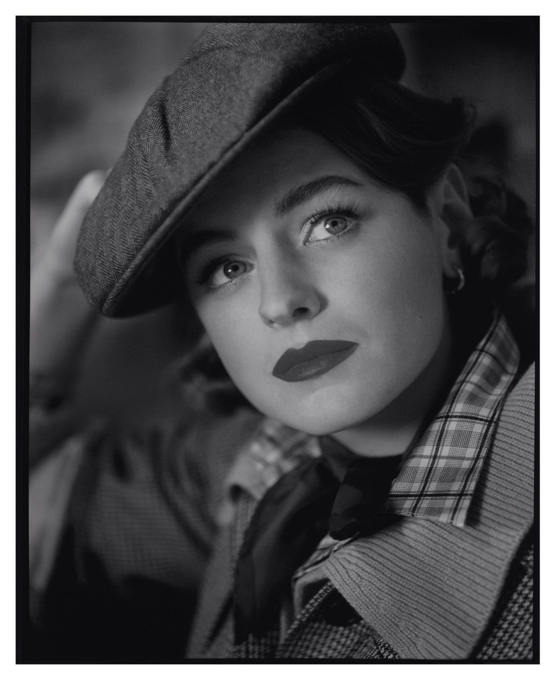 Monochromatic portrait of a woman wearing a newsboy cap, styled in a vintage fashion with red lipstick, captured in soft lighting.