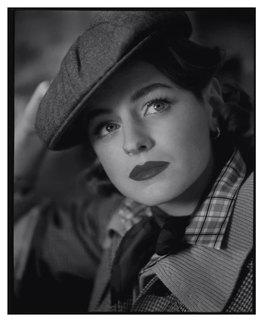 Monochromatic portrait of a woman wearing a newsboy cap, styled in a vintage fashion with red lipstick, captured in soft lighting.